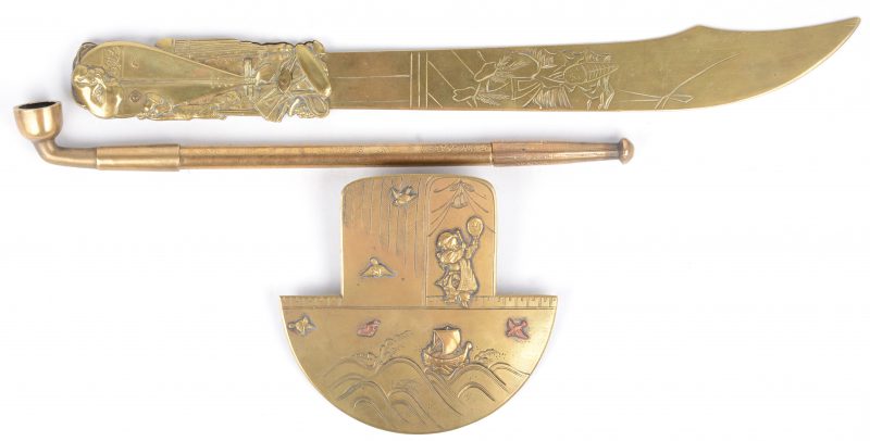 Drie Japanse voorwerpen van messing, bestaande uit een brievenopener, een helmmontuur en een pijp.