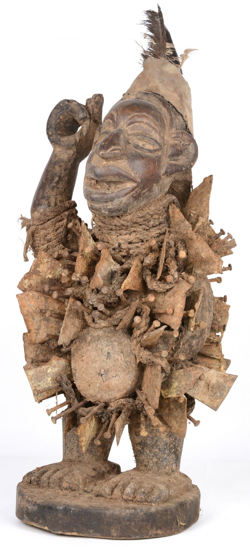Een Afrikaans fetisjbeeld van gebeeldhouwd hout met metalen platen en nagels en bovenaan getooid met veren.