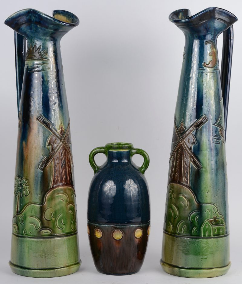 Twee hoge sierkruiken van Belgisch aardewerk, versierd met een reliëfdecor van een molenlandschap. We voegen er een gelijksoortig vaasje met twee oren aan toe.