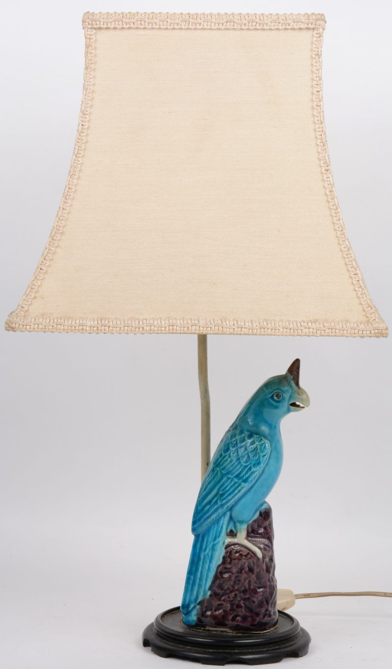Een lampje, opgebouwd uit een beeldje van meerkleurig aardewerk in de vorm van een vogel op een houten sokkeltje. Sokkeltje beschadigd.