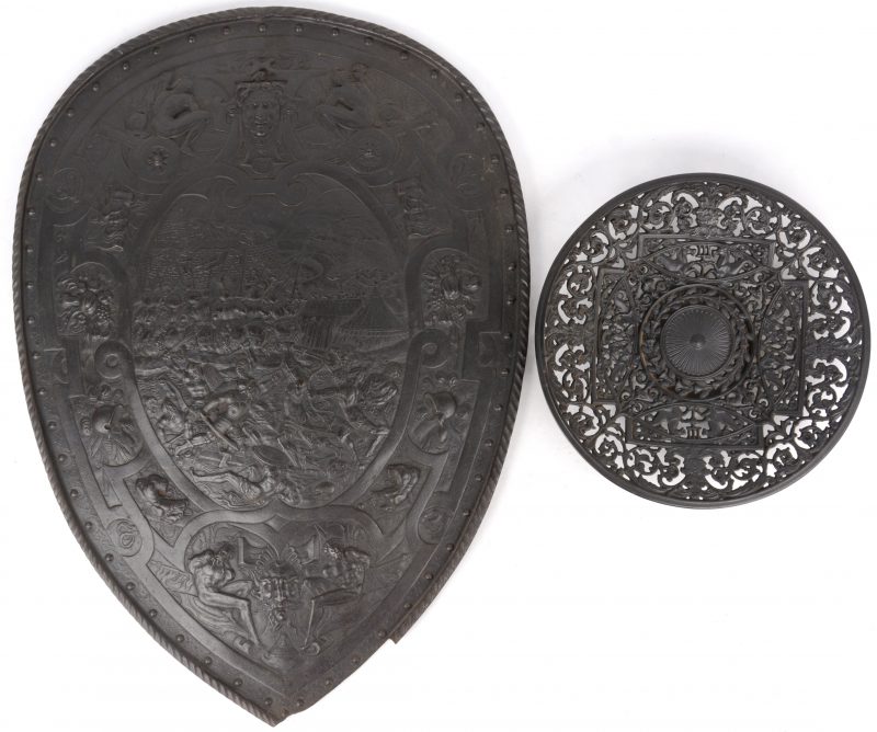 Een gietijzeren sierschild met een slagveldscène en heraldische symbolen in reliëf. Onderaan beschadigd. We voegen er een opengewerkte schaal, eveneens van gietijzer, aan toe.