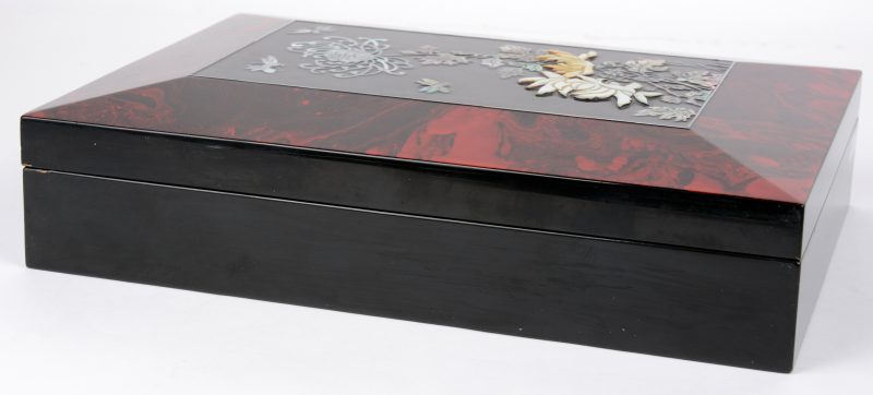 Een platte en rechthoekige Chinese doos, rondom in rode en zwarte lak, het deksel versierd met een decor van bloemen en insekten in parelmoer. Omstreeks 1930.