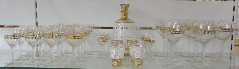 Een glasservies van kleurloos kristal met gouden versieringen, bestaande uit rodewijnglazen, wittewijnglazen, aperitiefglazen en borrelglazen op voet. Telkens 8 stuks. Evenals een karaf en vier borreltjes.