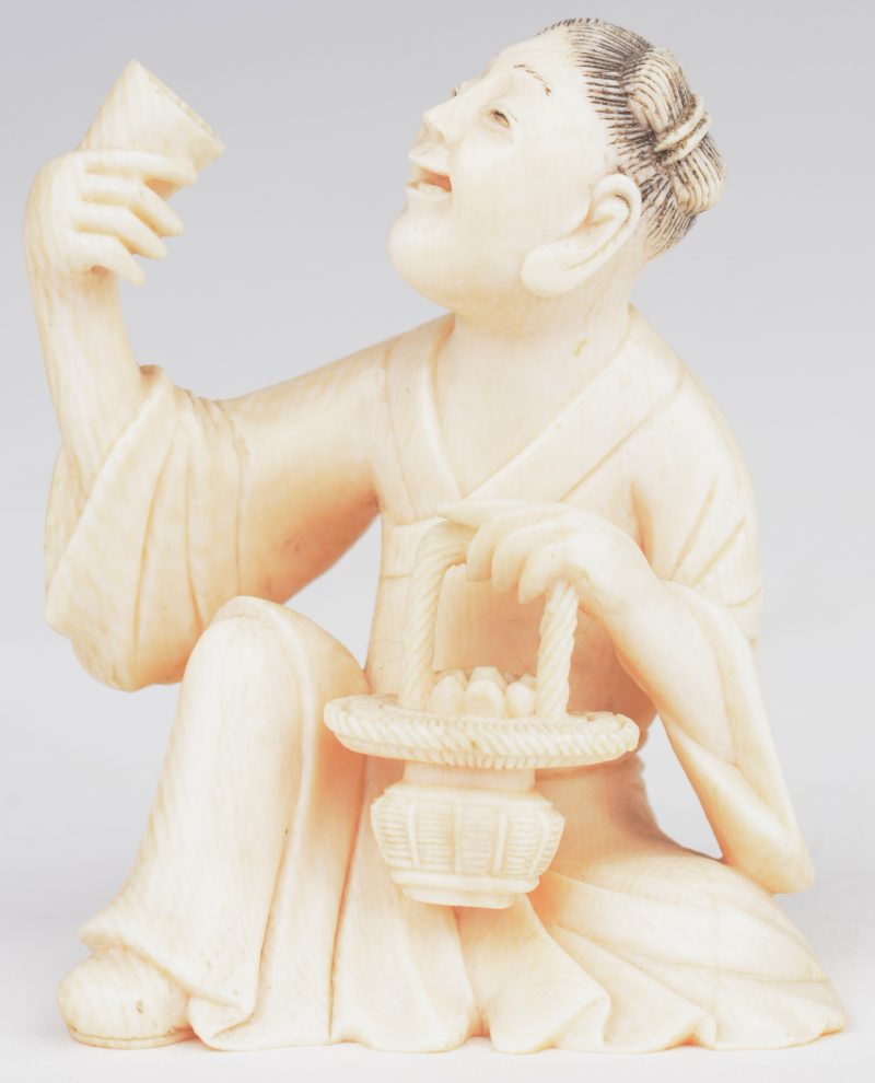 Okimono van gebeeldhouwd ivoor. Voorstelling van een zittende man met een beker en een mandje. Japans werk, begin XXste eeuw. Onderaan gemerkt.