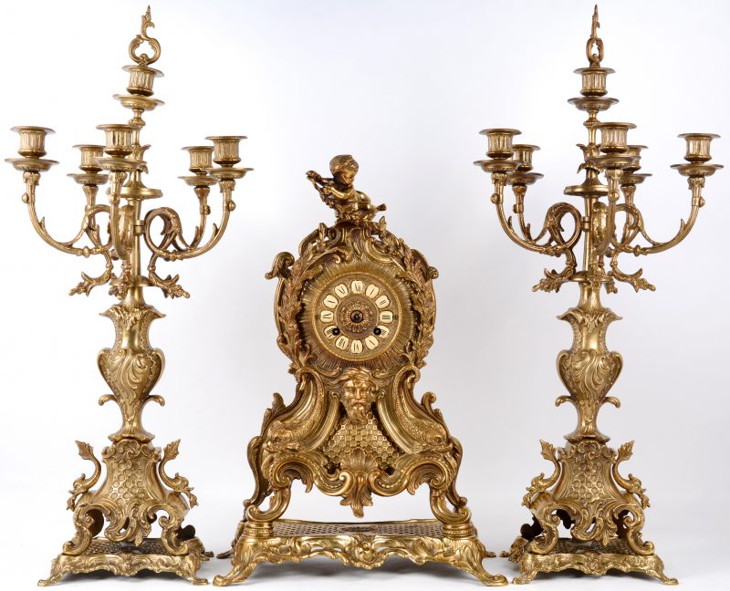 Een driedelig klokstel van verguld messing in Lodewijk XV-stijl, bestaande uit een pendule en twee grote kandelaars. De pendule versierd met barokke dolfijnen en bovenaan met een putto. Wijzers ontbreken.