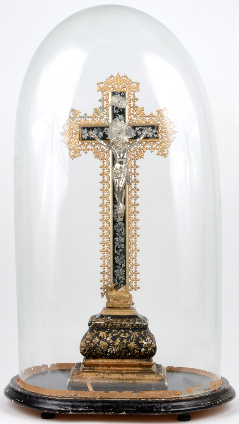 Een kruisbeeld van geploychromeerd en goudgepatineerd plaaster met een verzilverde corpus en een voorstelling van het Lam Gods aan de basis. Onder grote stolp.