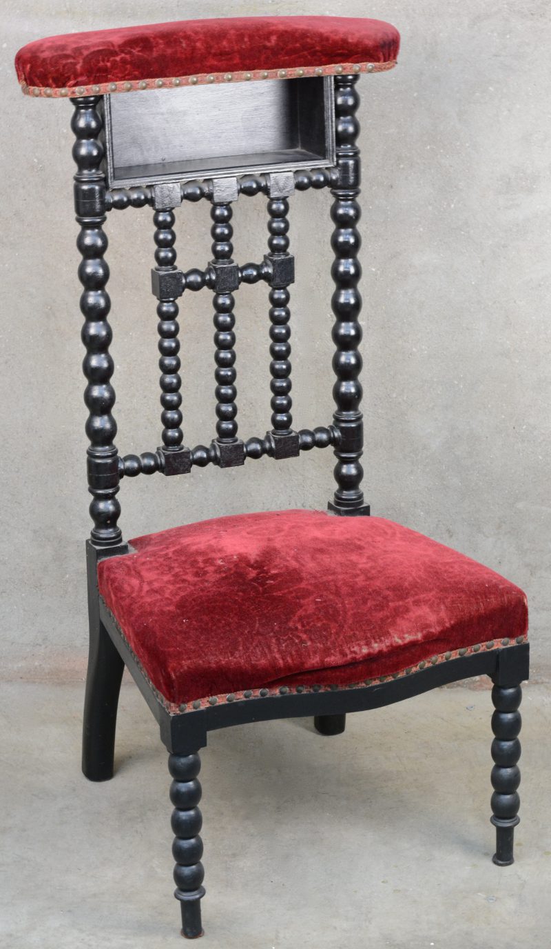 Een kerkstoel van zwartgepatineerd hout met gedraaide poten en spijlen en bekleed met rood fluweel.