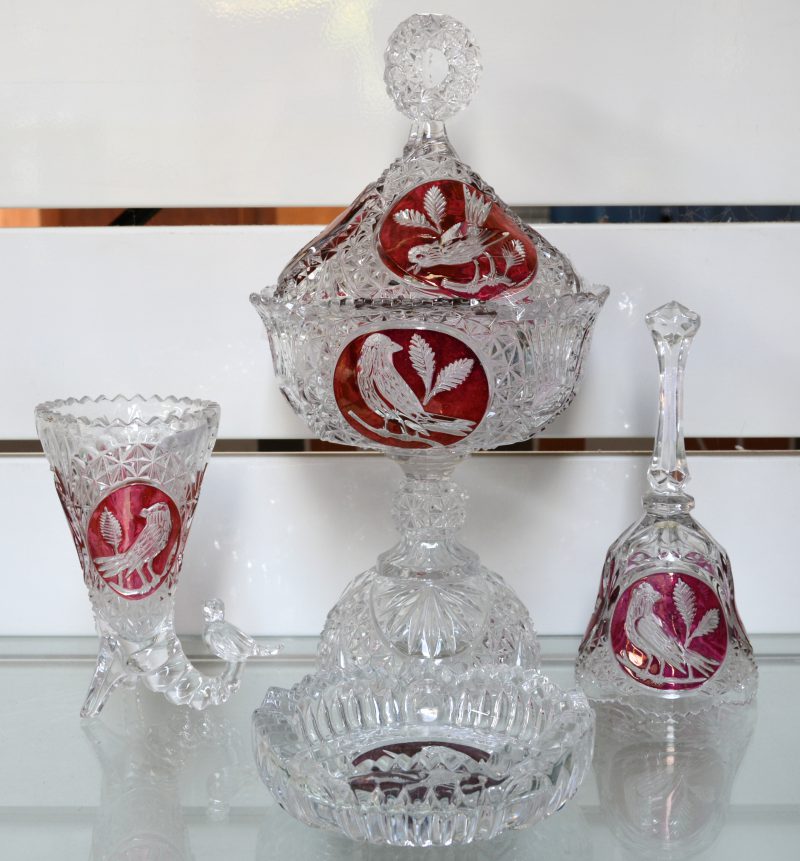 Een lot van vier voorwerpen in geslepen kristal met vogel in  rode cartouches . Bestaande uit een bonbonnière met deksel op voet, een bel, een asbak en een sierbekertje.