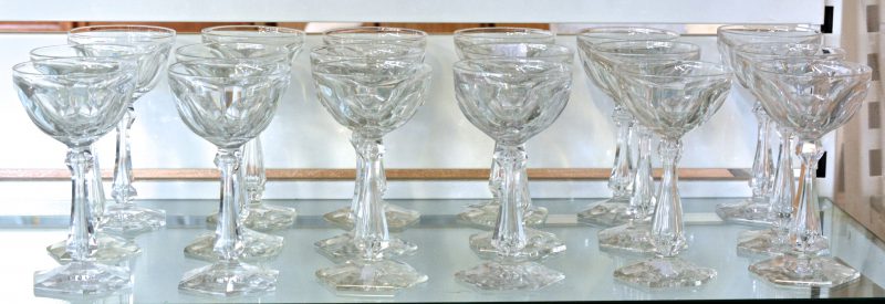 Een reeks van achttien glazen van geslepen kleurloos kristal op zeshoekige voet. Enkele glazen met kleine schilfer aan de voet.