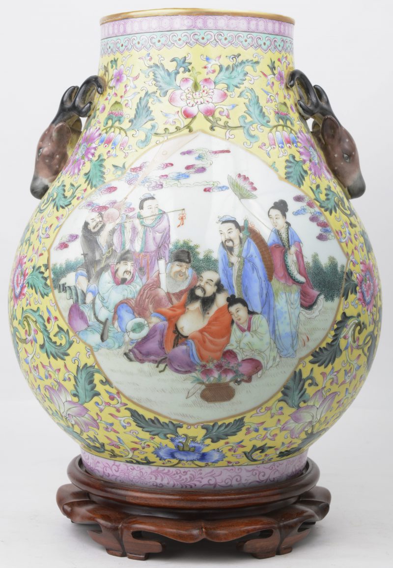 Een vaas van Chinees porselein met een decor van personages in cartouches op een gele fond met bloemen en versierd met twee hertenkoppen in reliëf. Met sokkel.