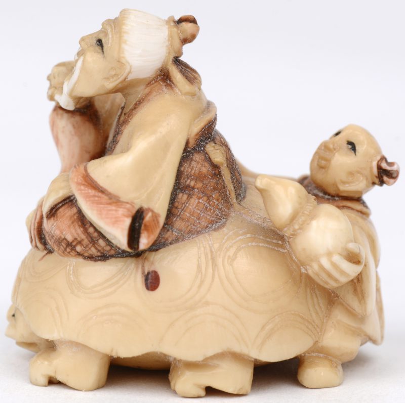 “Man met jongen op schildpad”. Een netsuke van gesculpteerd ivoor met gepolychromeerde details. Onderaan gesigneerd. Eerste kwart XXe eeuw.