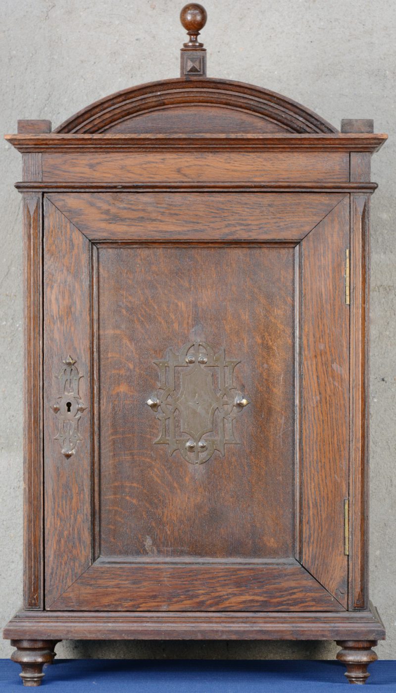 Een klein eikenhouten wandkabinetje met één deurtje met koperen versiering en met twee kleine laden binnenin.