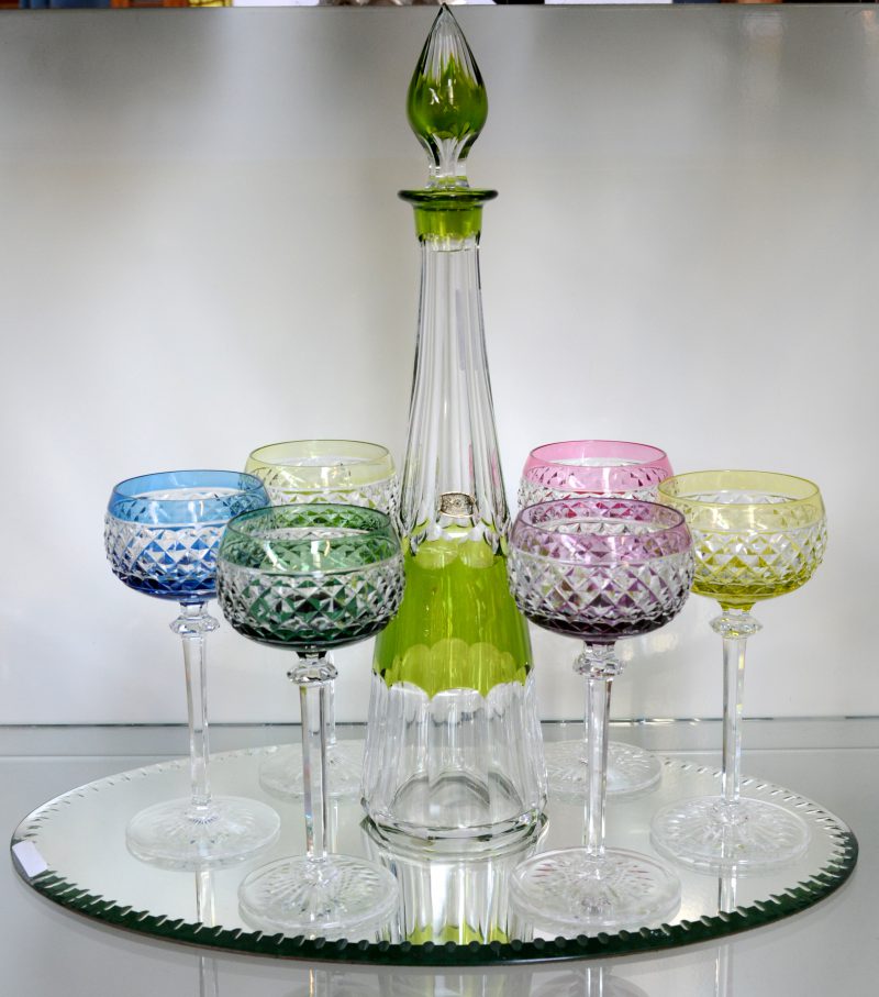 Een karaf en zes bijpassende glazen van geslepen kristal in diverse kleuren. Op presenteerspiegel. Gemerkt.