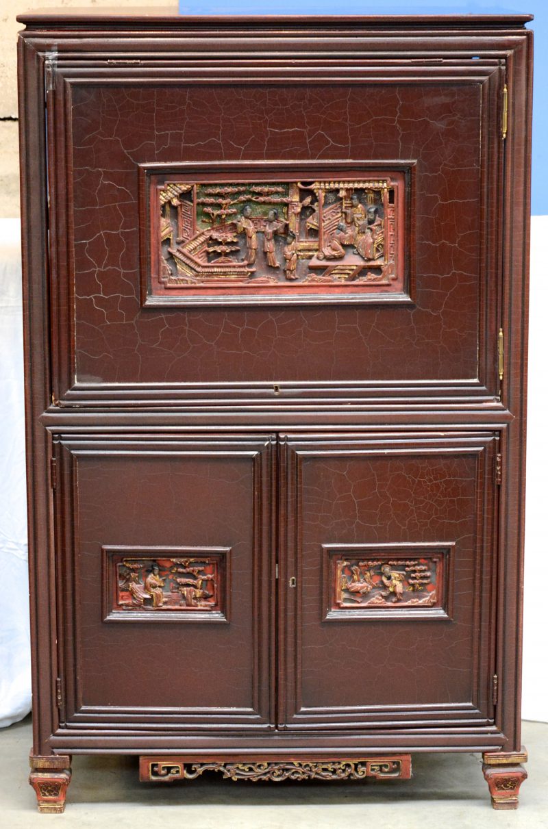 Een Chinees televisiemeubel van roodgelakt hout met twee kleine paneeldeuren onder een grote, telkens versierd met een uitgestoken decor van personages.