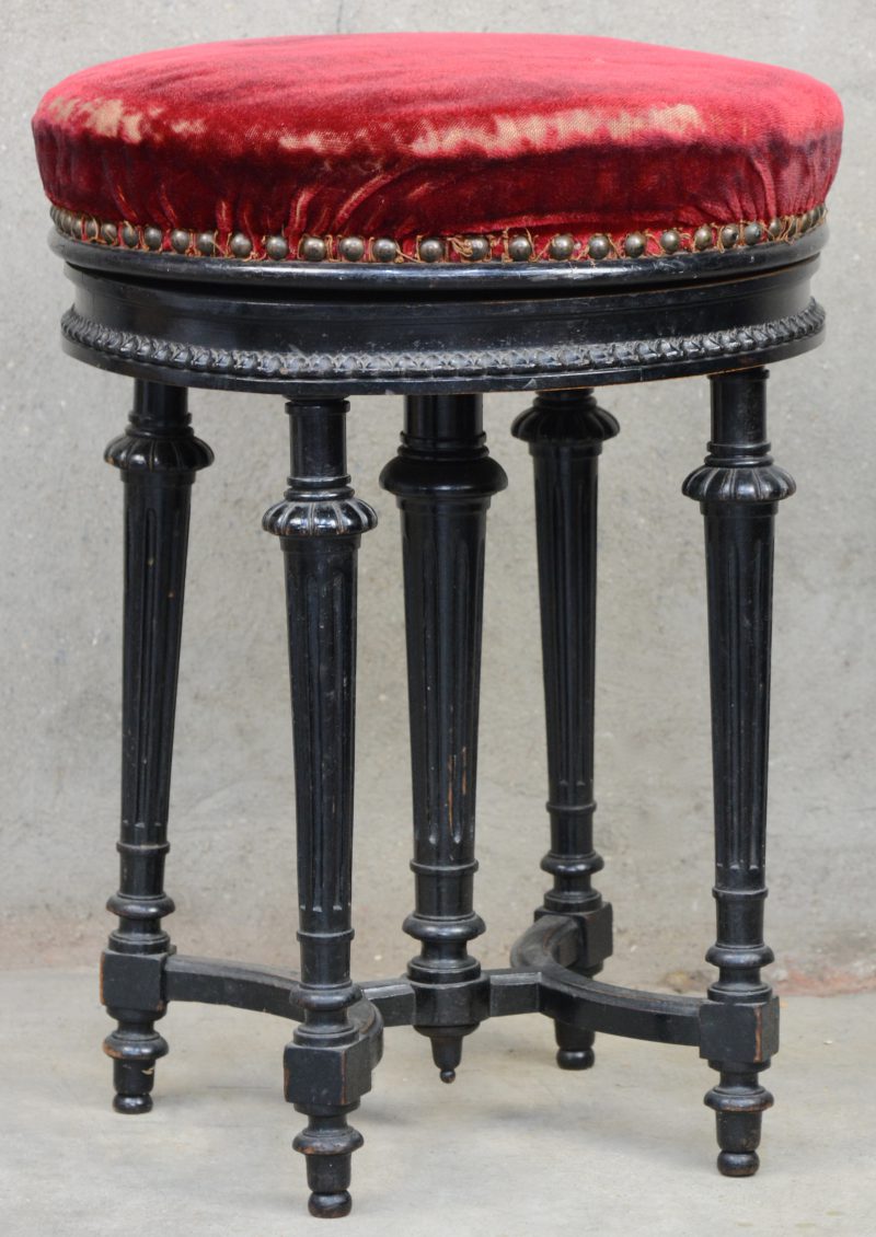 Een antiek pianostoeltje van zwartgelakt hout in Lodewijk XVI-stijl met roterende zit, bekleed met rood fluweel.