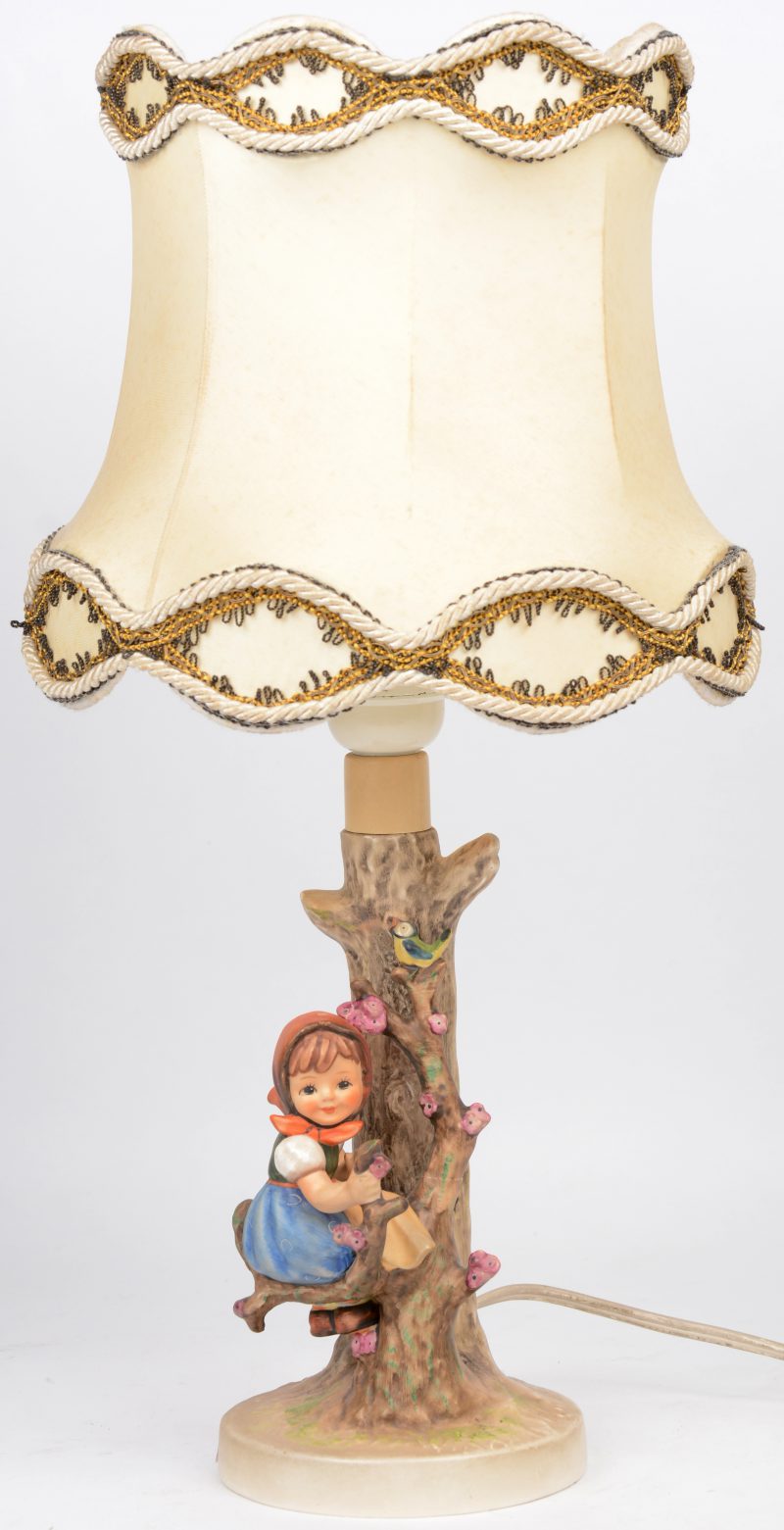 Een lampenvoet van meerkleurig porselein in de vorm van een meisje aan een boomstam. Uit de reeks van M.I. Hummel.