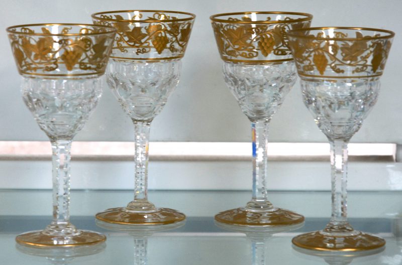 Een reeks van vier kleine glaasjes van kleurloos kristal op voet, versierd met een verguld decor van druivenranken.