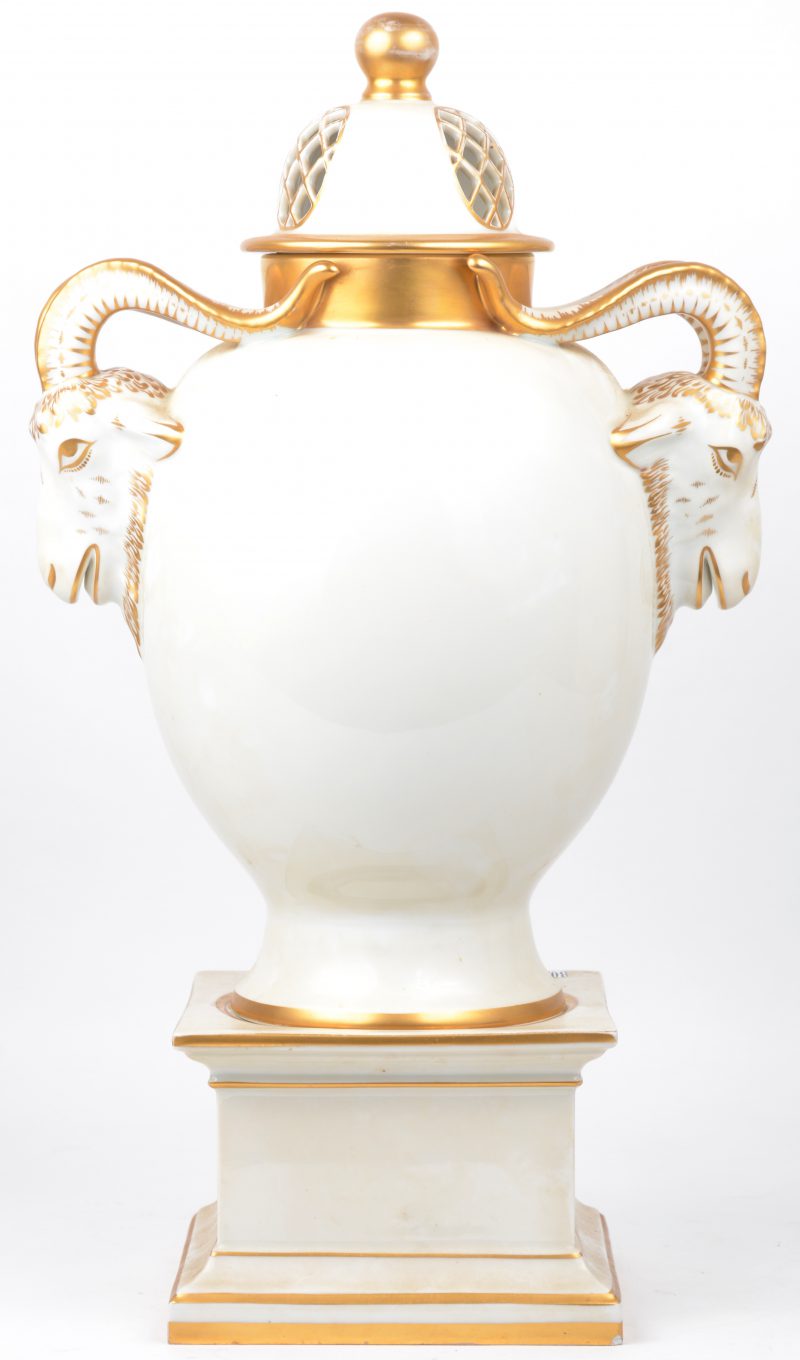 Een dekselvaas van met goud versierd Brussels porselein met handvatten in de vorm van ramskoppen en op een piëdestal.