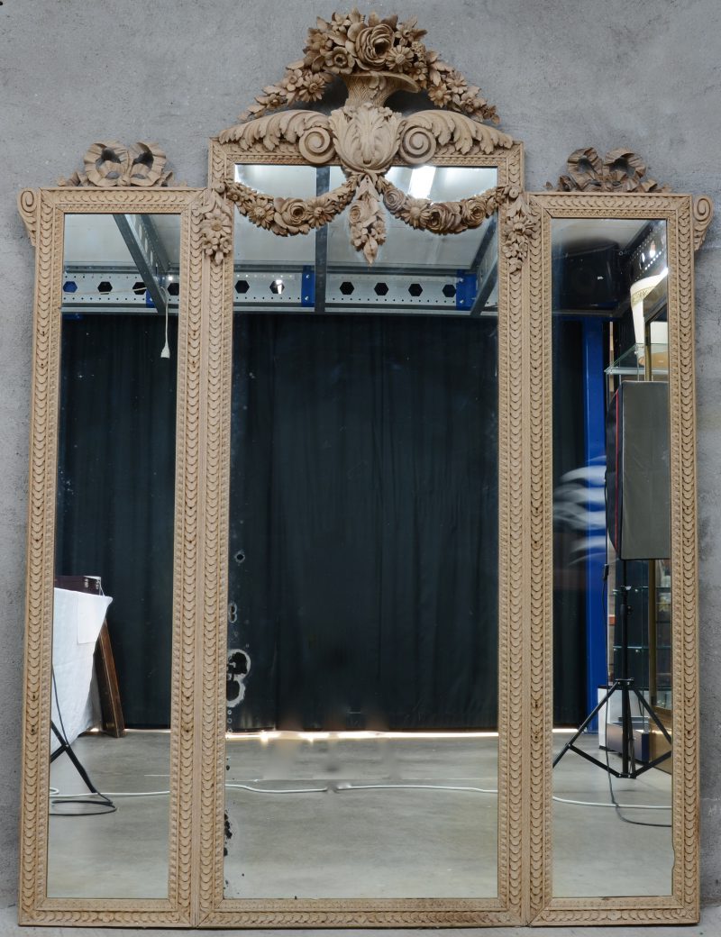 Een grote driedelige schouwspiegel van afgeloogd hout in Lodewijk XVI-stijl, versierd met bloemenguirlandes en strikken.