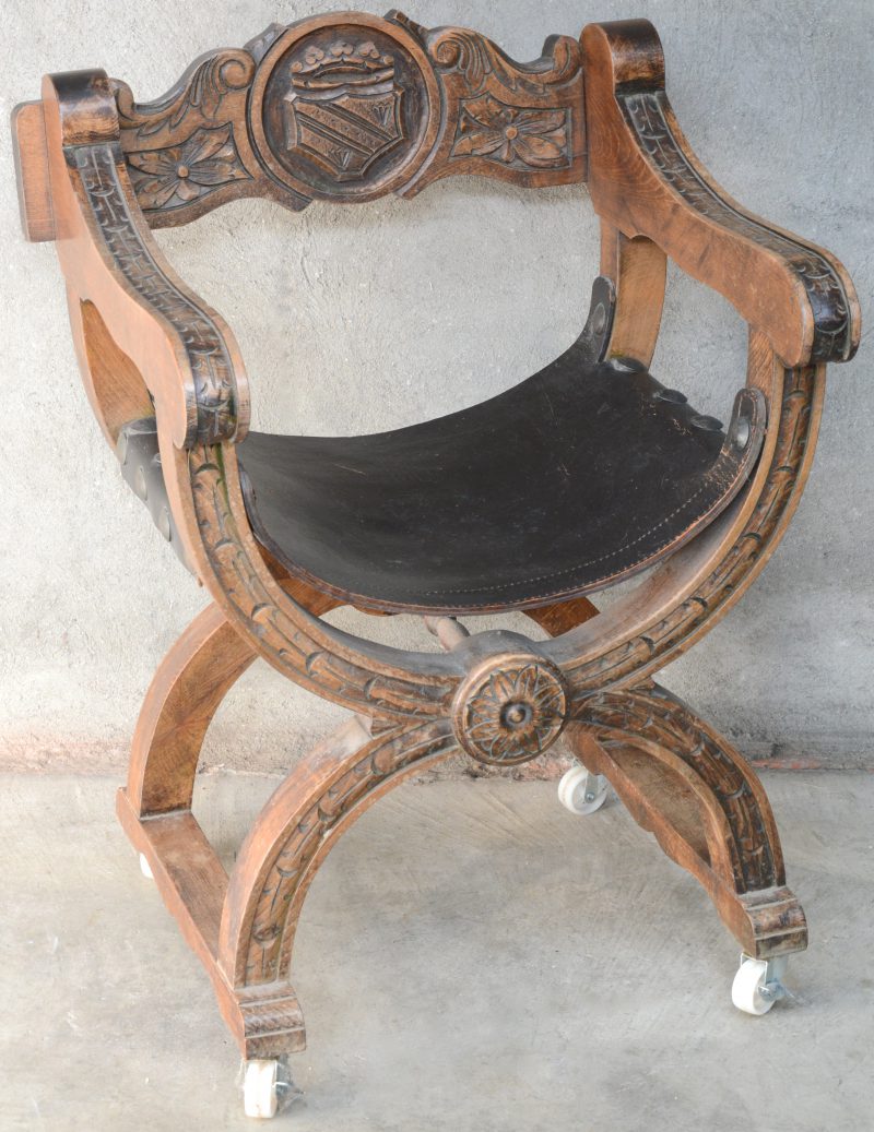 Een dagobertstoel van gebeeldhouwd eikenhout met een schild in de rug en met een lederen zit. Op wieltjes.