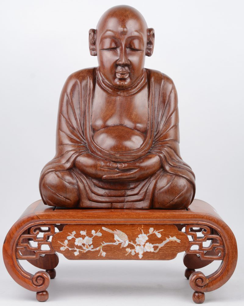 Een zittende Boeddha van gebeeldhouwd hout op een voetstukje, versierd met ingelegde tekens van parelmoer.