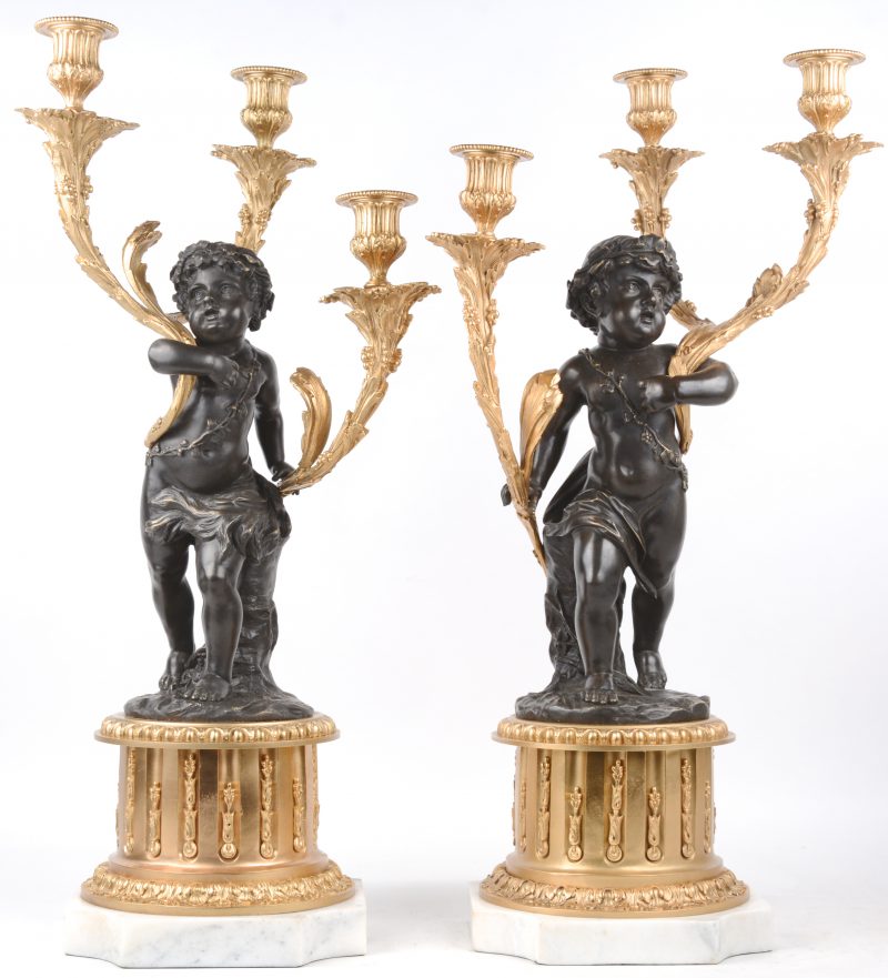 Een paar kandelaars van brons met telkens drie vergulde lichtpunten, gedragen door een putto en op een voetstuk van wit marmer.