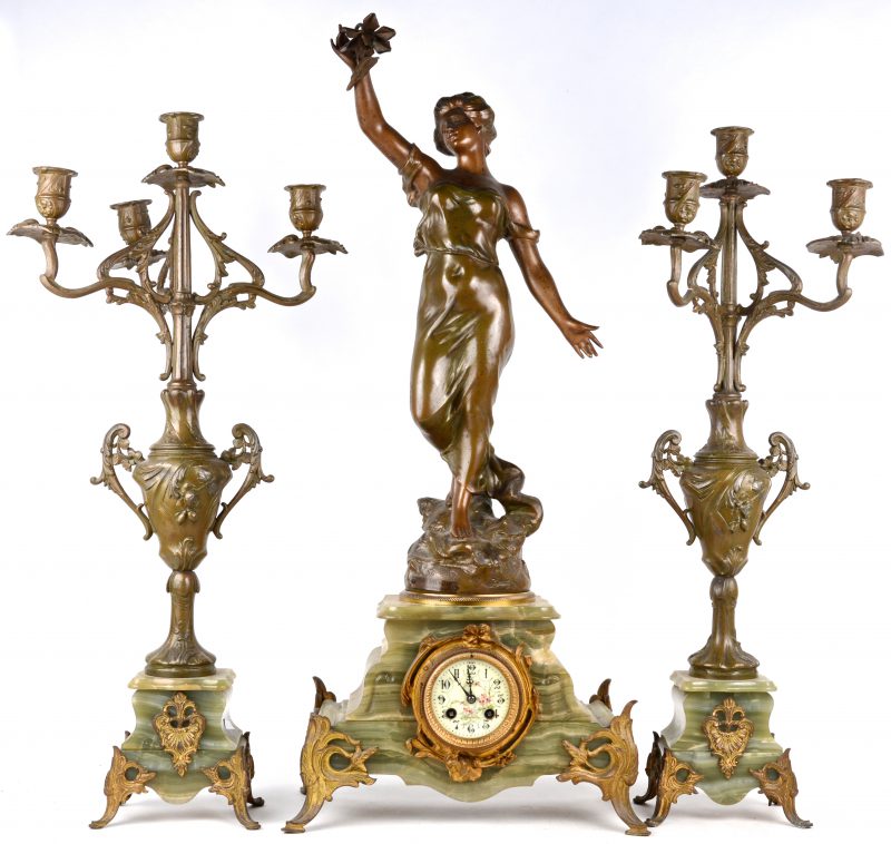 Een driedelig klokstel van zamak en onyx. Pendule versierd met een beeld getiteld “Brise du Soir par Stéphane”. Twee kandelaars waarvan één met een ontbrekende lichtarm. Omstreeks 1900.