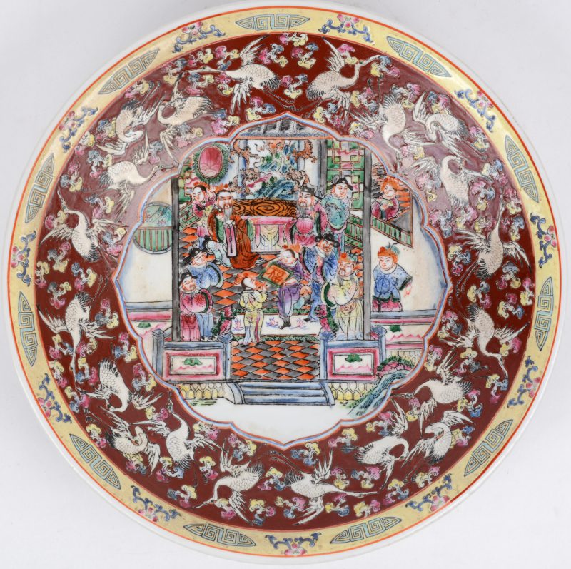 Een groot bord van Chinees porselein met een decor van personages in het plat en kraanvogels op rode achtergrond op de vleugel. Onderaan gemerkt.