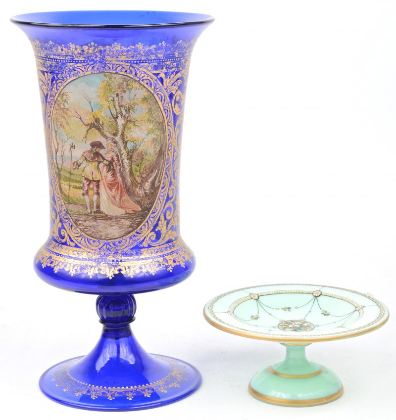 Een siervaas van blauw glas met vergulde versieringen en een handgeschilderde pastorale scène. We voegen er een opaalglazen schaaltje op voet met emailversieringen aan toe.