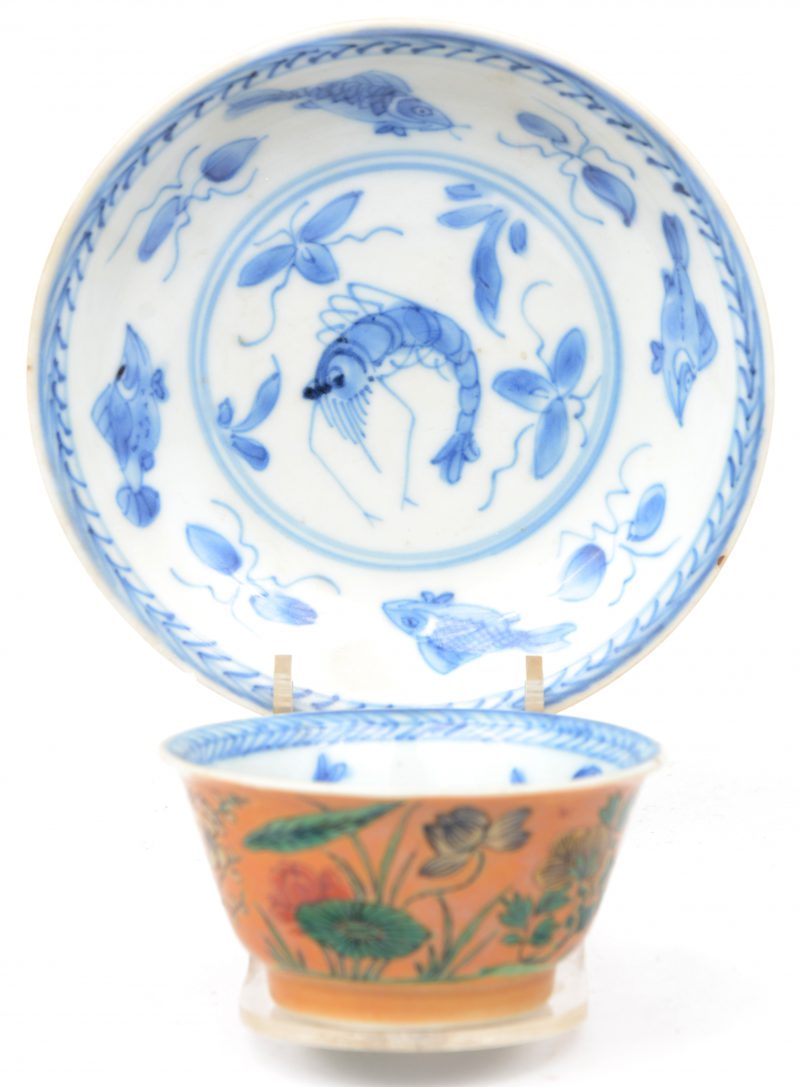 Een theekommetje met onderbordje met een blauw op wit decor van vissen en een kreeft binnenin en aan de buitenlant meerkleurige pioenen. Onderaan gemerkt. XIXe eeuw. Kleine schilfer aan het bordje.