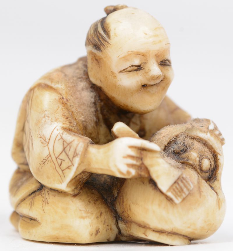 Een antieke Japanse netsuke van gesculpteerd ivoor ion de vorm van een zittende man die een masker maakt. Onderaan gesigneerd.