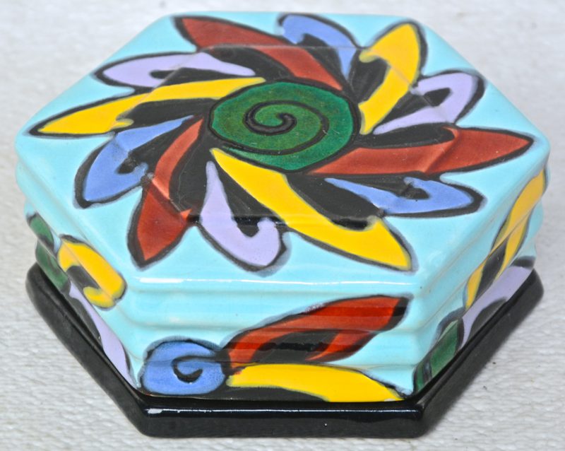 Een zeshoekig art deco dekseldoosje van aardewerk met een meerkleurig decor. Onderaan gemerkt. Schilfer aan de binnenkant.