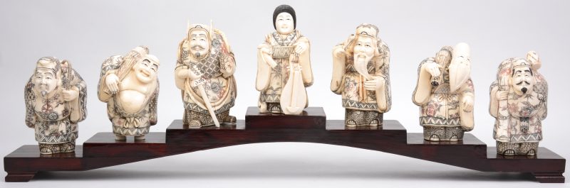 Een reeks van zeven beelden van gebeeldhouwd en gepolychromeerd ivoor op hout die de zeven Japanse geluksgoden (Shichi Fukujin) voorstellen. Onderaan gesigneerd. Op houten display.