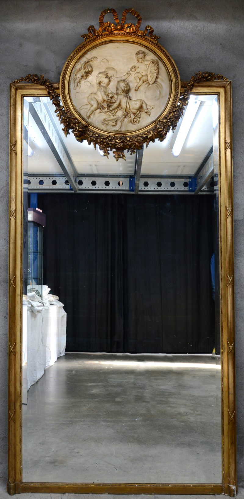 Een grote schouwspiegel in Lodewijk XVI-stijl met verguld houten lijst en met een beigegepatineerd bas-reliëf met een voorstelling met putti. Begin XXe eeuw.