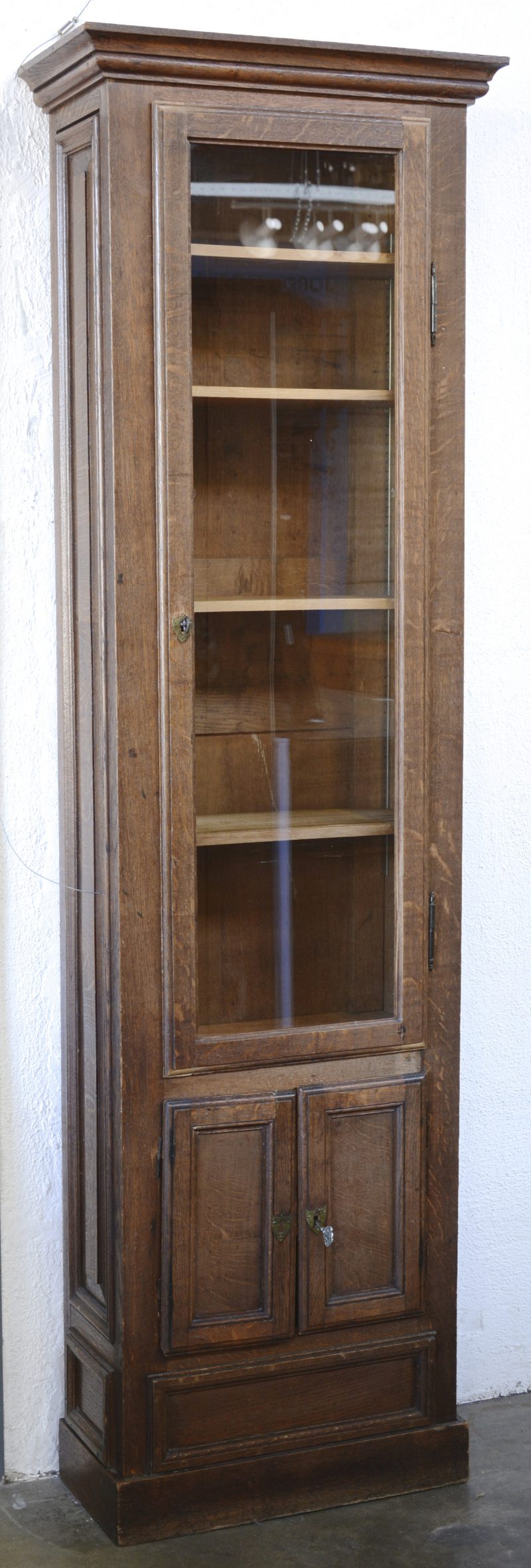 Een hoge eikenhouten vitrinekast met twee paneeldeurtjes onder de glazen deur.