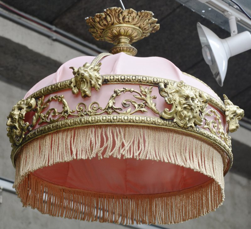 Een bronzen hanglamp, versierd met leeuwen- en saterkoppen en met roze stoffen kap.