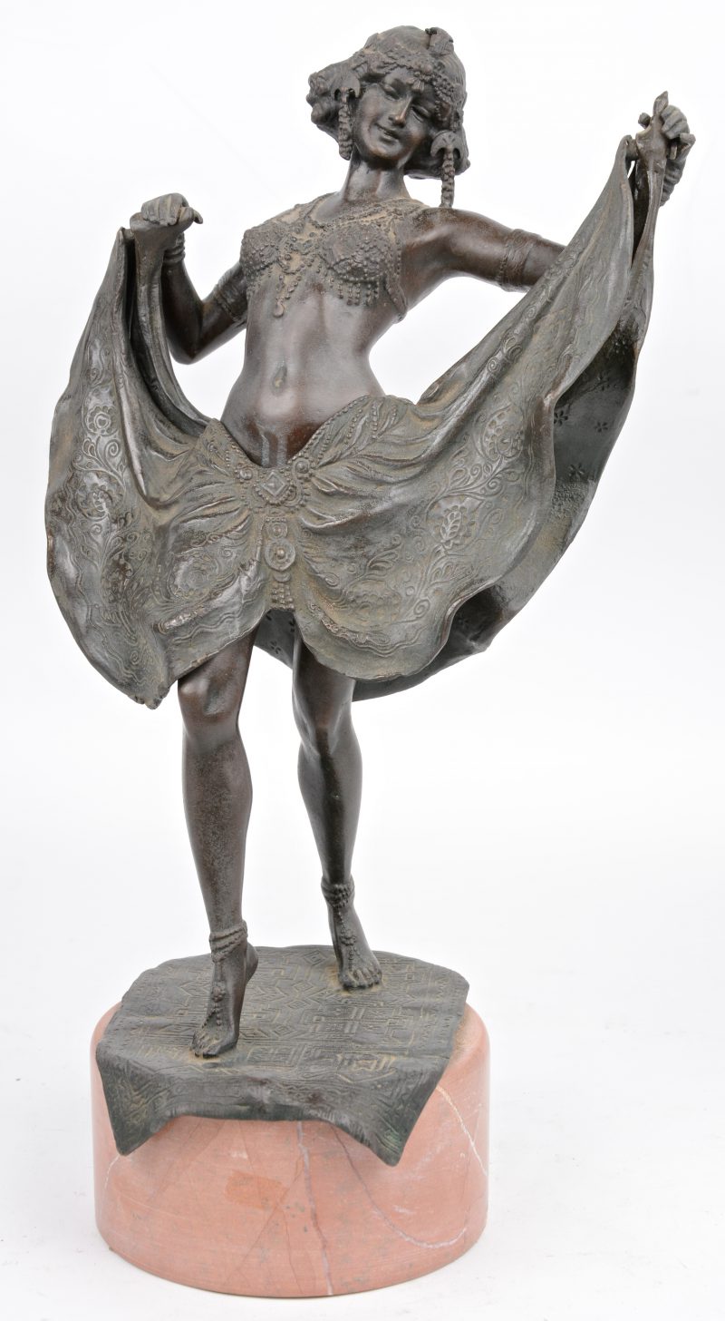 Een danseres in klederdracht uit de jaren ‘20 van donkergepatineerd brons met een opklapbaar kleedje. Op voetstuk van roze marmer.