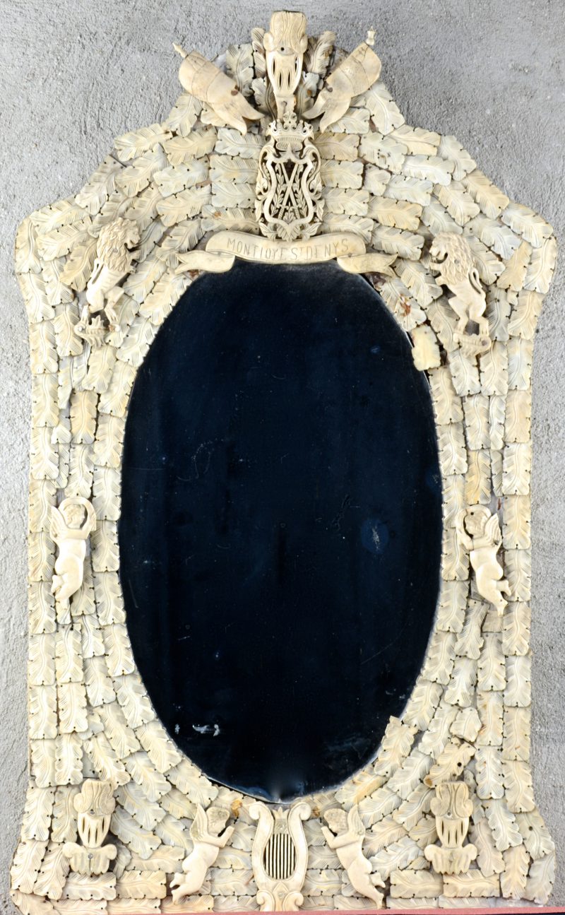 Een antieke spiegel, bekleed met gesculpteerd benen plaatjes met Engeltjes en leeuwen en centraal het wapenschild van het Franse koninkrijk en de spreuk “Montjoy Saint Denis”. Enkele manco’s en kleine beschadigingen.