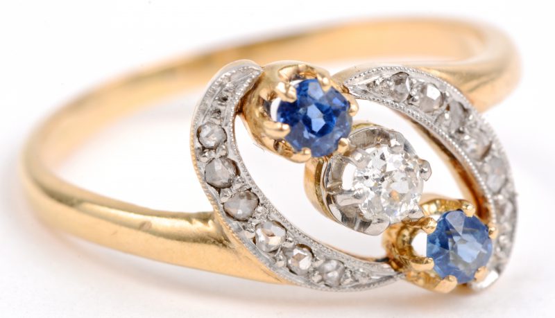 Een 18 karaats wit en geel gouden ring bezet diamanten oude slijp met een gezamenlijk gewicht van ± 0,12 ct. een centrale briljant van ± 0,15 ct. en twee saffieren met een gezamenlijk gewicht van ± 0,10 ct.