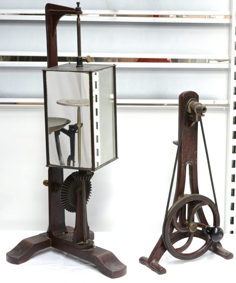 Een manueel aandrijfwiel en een roterende balkvormige spiegel. Beide op gietijzeren voet. Omstreeks 1900.