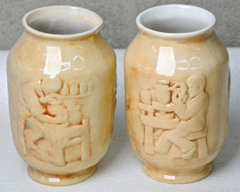 Twee bruingeglazuurde aardewerken vaasjes met een reliëfdecor van pottebakkerijtaferelen. Onderaan gemerkt. Gaaf.