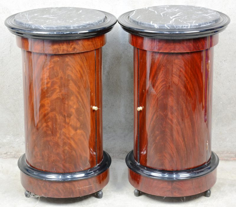 Een paar ronde bijzetkastjes van gefineerd mahoniehout met één lade en een zwart marmeren blad.