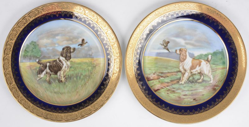 Een paar borden van Frans porselein met een gehoogd meerkleurig decor van jachthonden in het plat, met een kobaltblauwe vleugel en met vergulde rand.