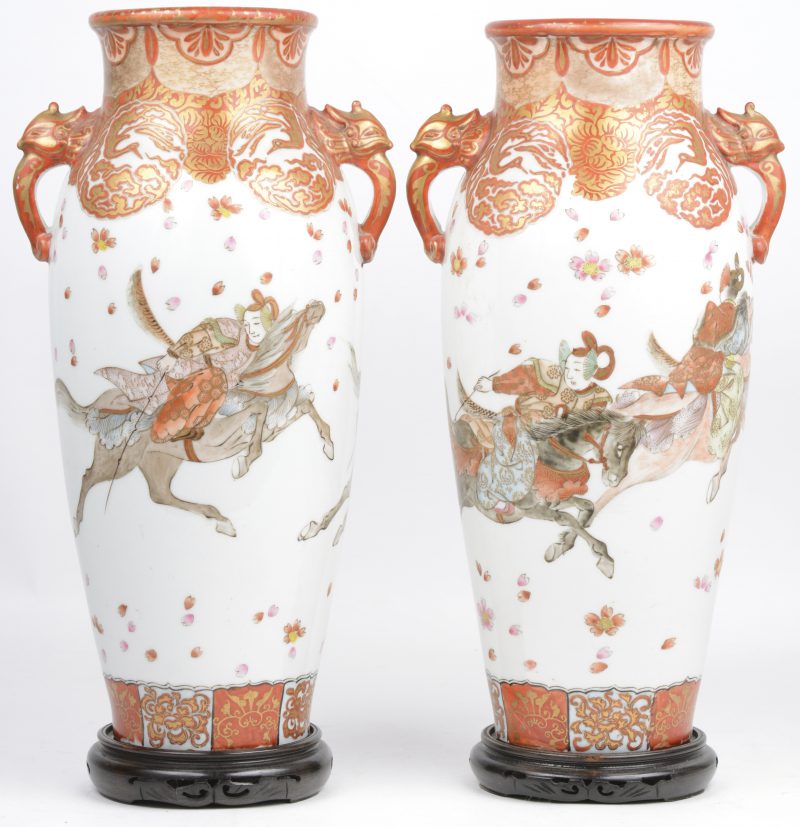 Een paar vazen van Japans porselein met decor van krijgers op roestrode en witte fond. Oren in de vorm van fabeldieren. Onderaan Kutani gemerkt, ostreeks 1930. Met houten sokkeltjes.
