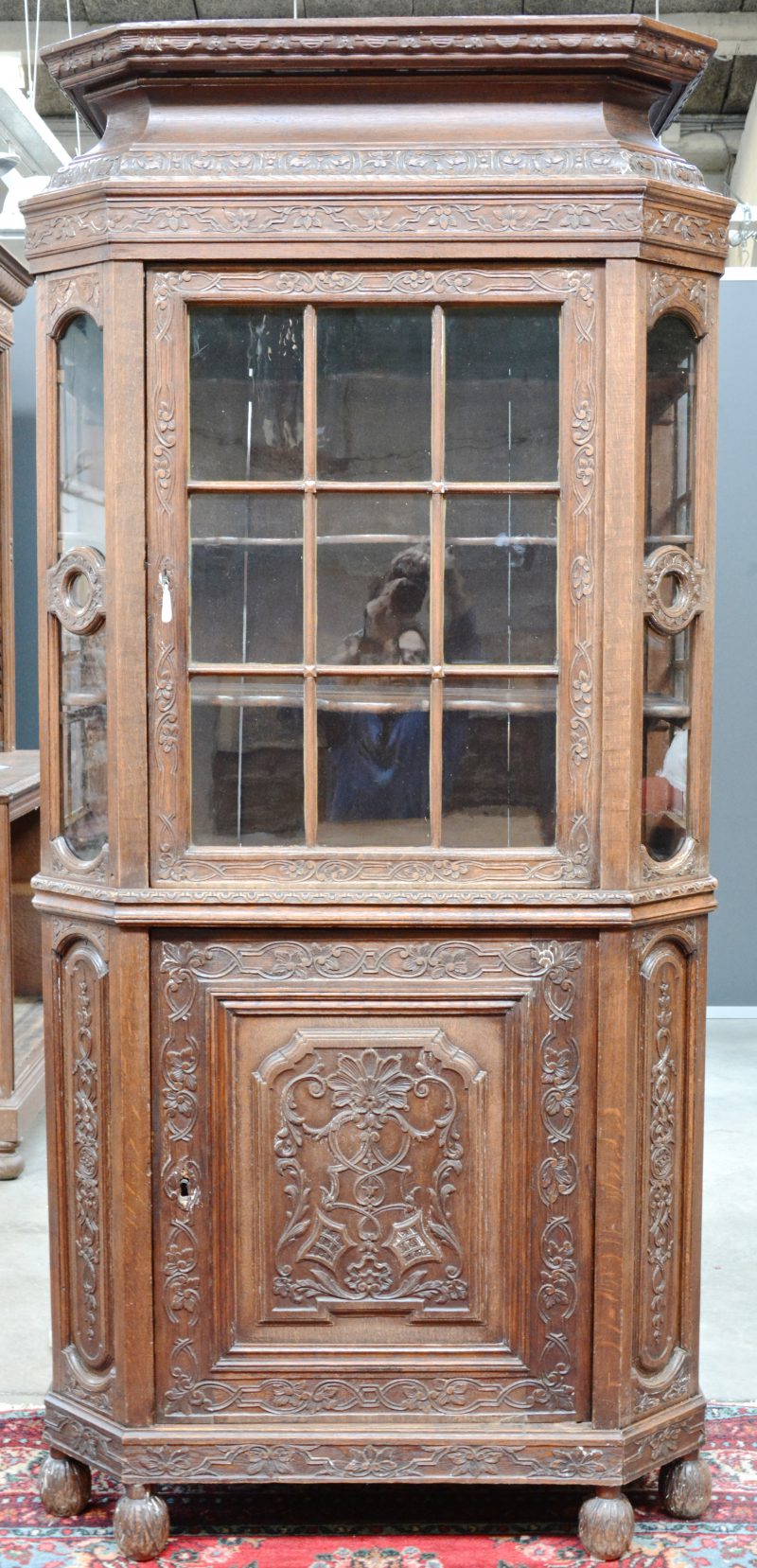 Een smalle vitrinekast van gebeeldhouwd eikenhout met een beglaasde deur boven een paneeldeur. Schuine stijlen, lancierskap, rijkelijk versierd met Régencemotieven. Frankrijk, medio XIXde eeuw.