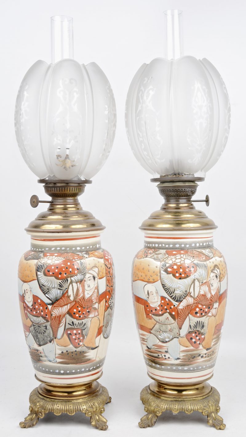 Een paar olielampen van koper en Japans aardewerk met decor van personages. Met lampglazen en lampenkappen van glas.