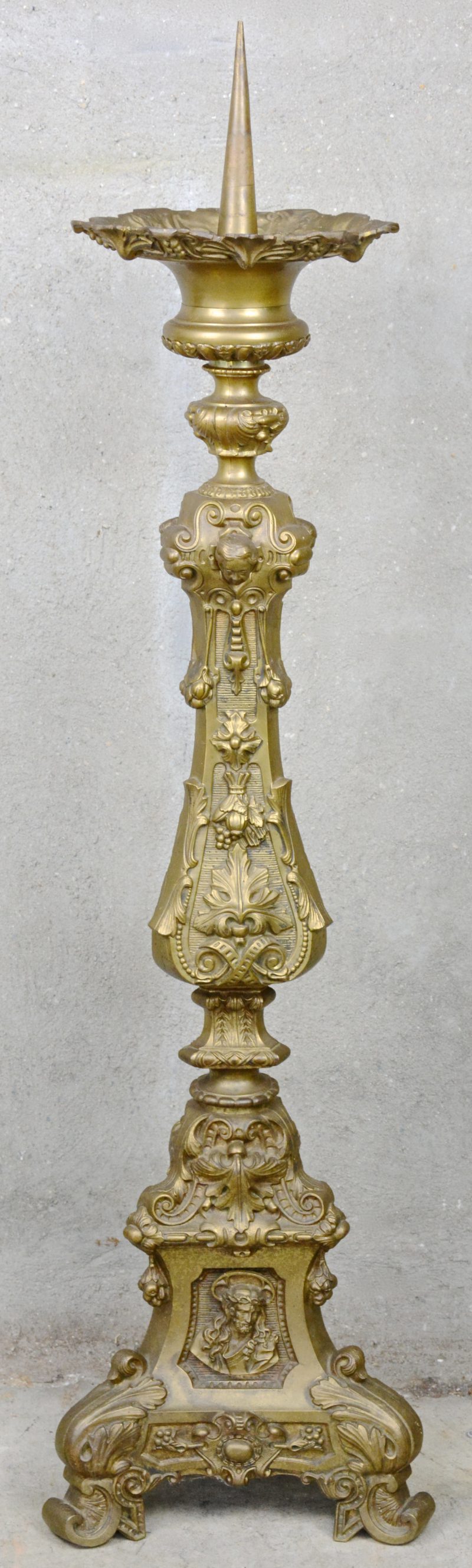 Een grote barokke kerkkandelaar van gedreven messing. Driehoekige basis met Heilige Harten, uitgebreide bladmotieven op de schacht, bovenaan drie engelenkopjes in reliëf.