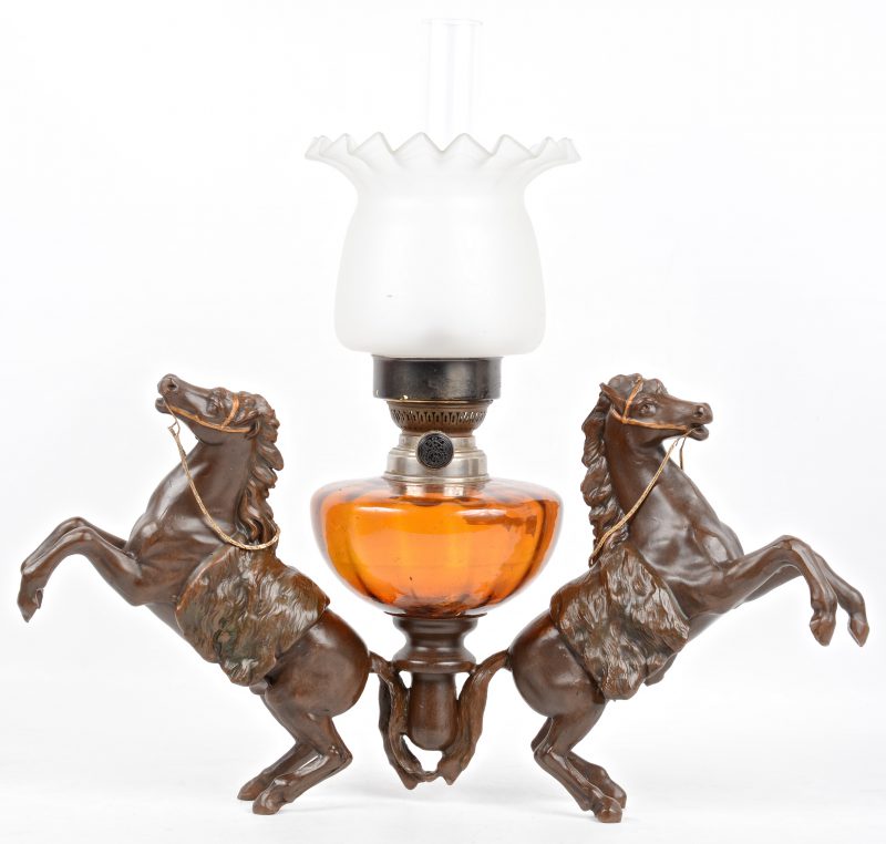 Een olielamp met oranje glazen recipiënt, gedragen door twee steigerende paarden van zamak.