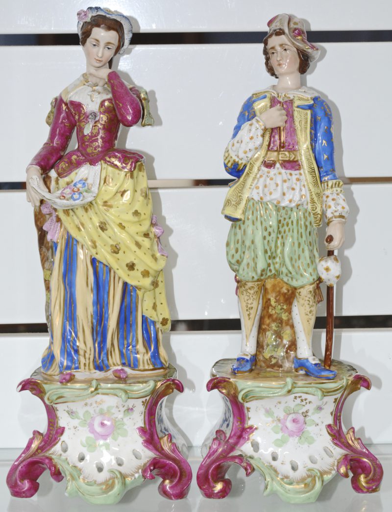 Een paar gepolychromeerde porseleinen beeldjes van een man en een vrouw. Weelderig gekleurd. Schade aan één voet.