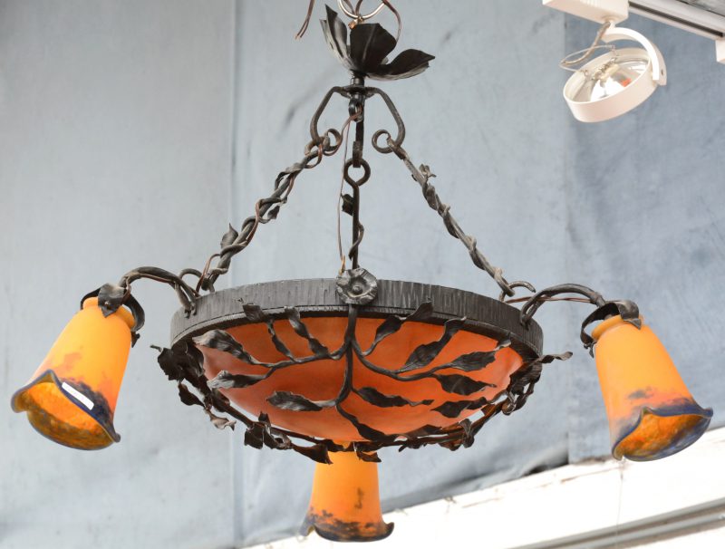 Een ronde hallantaarn bestaande uit een oranje glaspasta coupe gevat in smeedijzer met een bloem-en bladmotief. Drie externe lichtarmen met glaspasta lampenkappen gemerkt Degué en met verlichting in de coupe.
